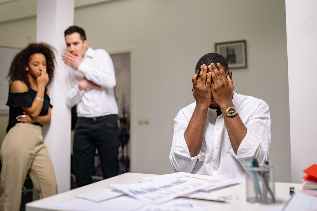 Stressed employee at a desk covered with pay documents while colleagues react behind him
