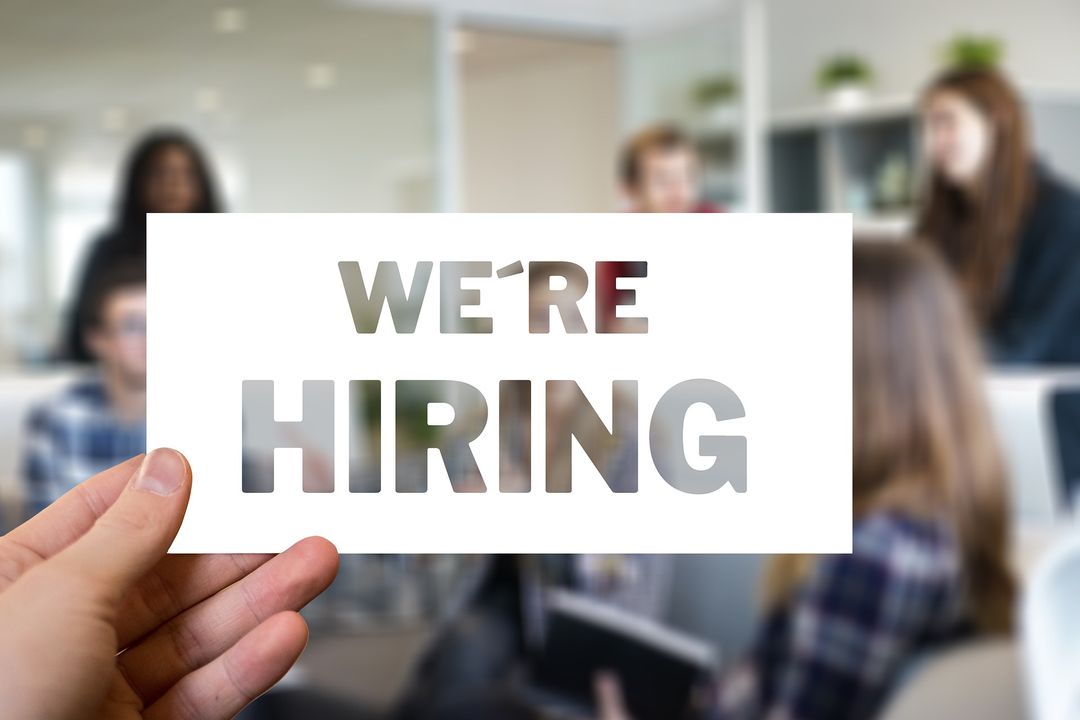 Hand holding a we are hiring sign in front of a recruiting team meeting a candidate in an office